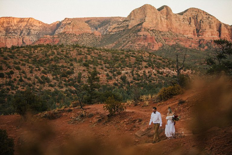 Bride in a wedding dress walking with her new husband in Sedona, arizona after their Cathedral rock elopement. Red rocks of Sedona in the background.